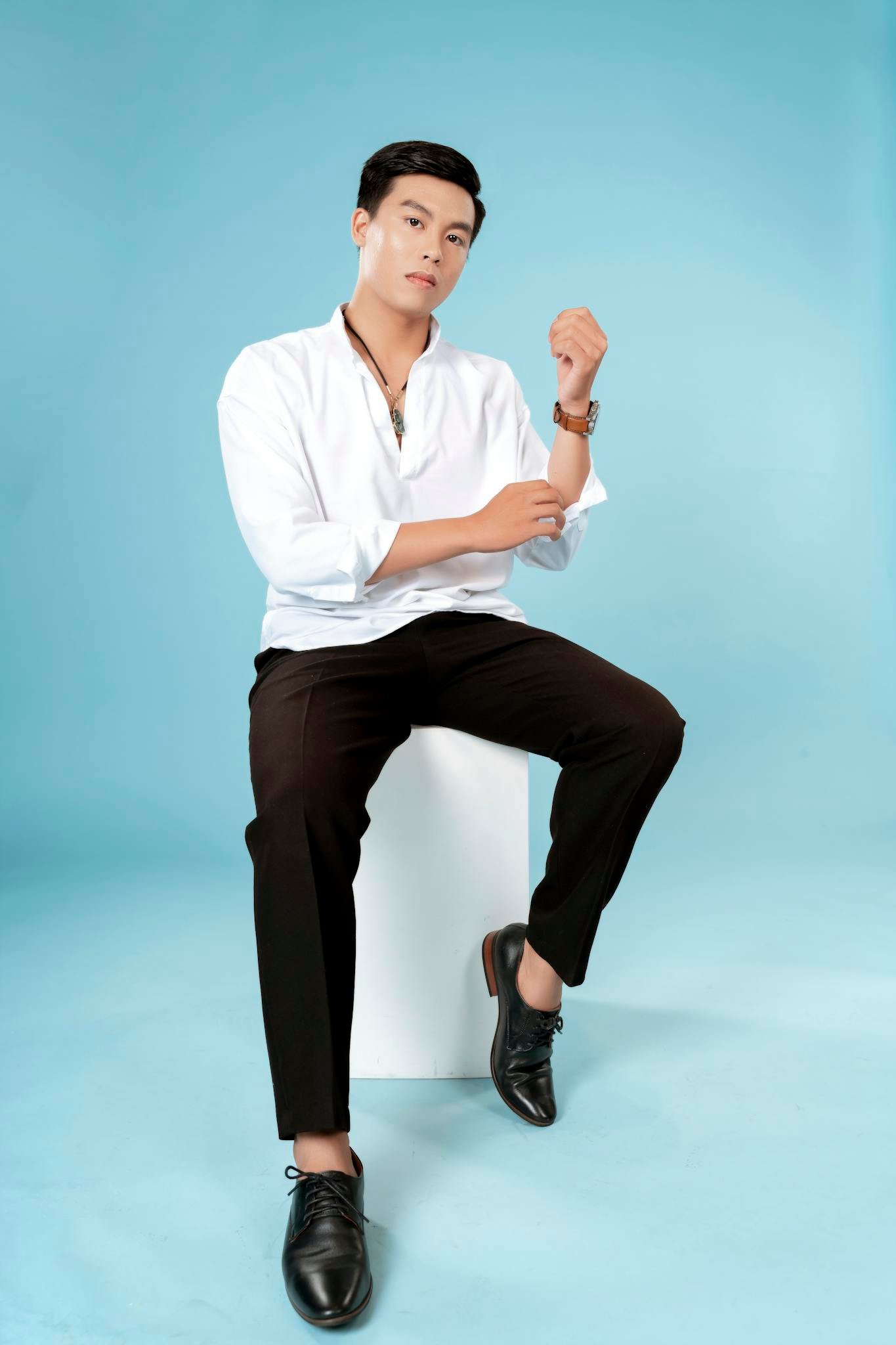 Confident man in a modern studio setting wearing a white shirt and black pants, seated against a blue background.