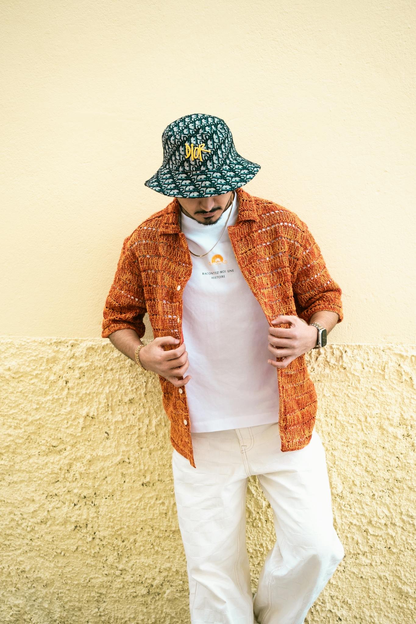 Fashionable man in Adana embraces casual style with orange shirt and patterned hat against a textured wall.